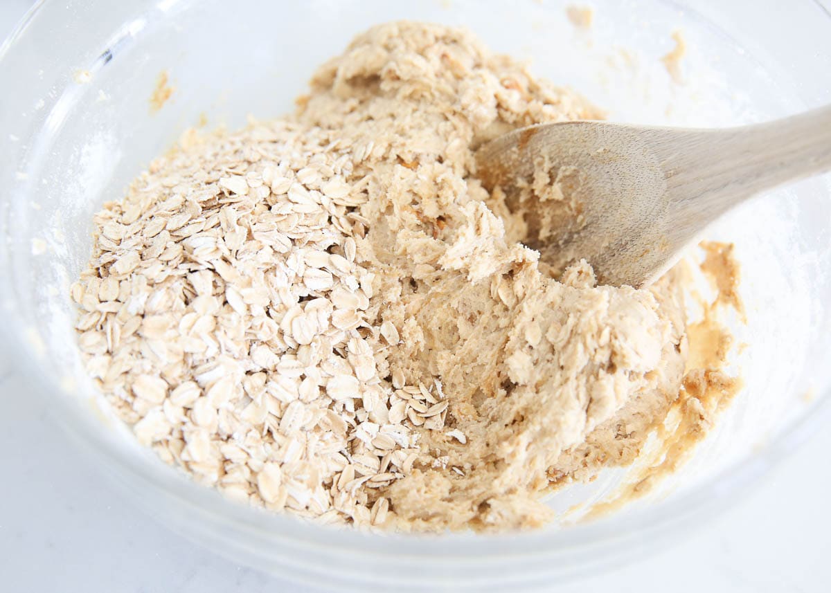 Using a wooden spoon to stir in oats to cookie batter in a bowl.