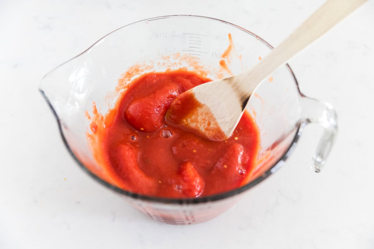 Crushing whole tomatoes in a measuring cup with spoon.