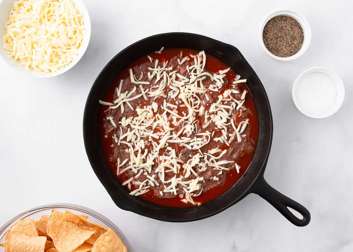 Refried beans spread over enchilada sauce in a skillet with cheese.