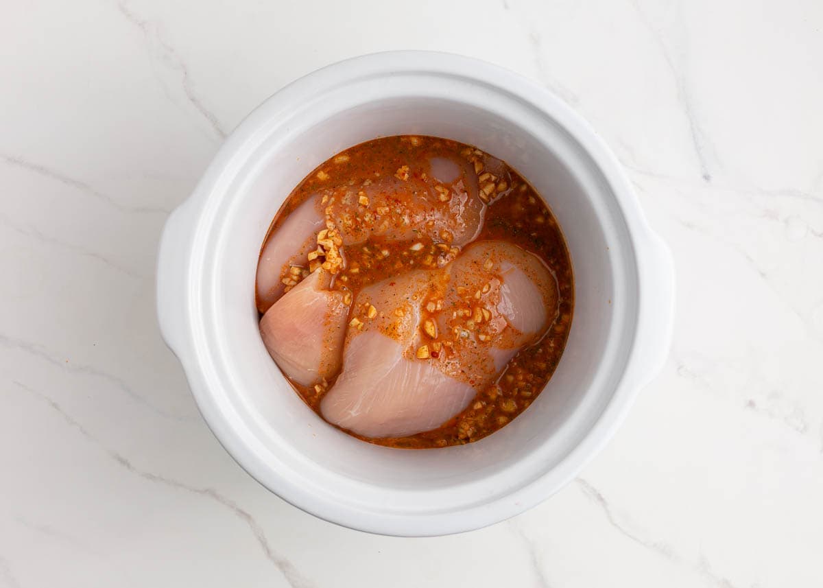 Chicken breast in a slow cooker with seasoning and liquid poured on top.