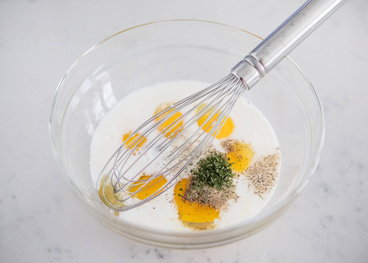 Eggs in a bowl with milk, seasonings and a whisk.