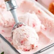 Scooping homemade strawberry ice cream out of a glass container.