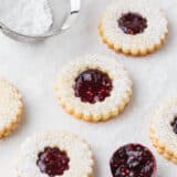 Linzer raspberry cookies on the counter with jam in a spoon and powdered sugar in a sifter.