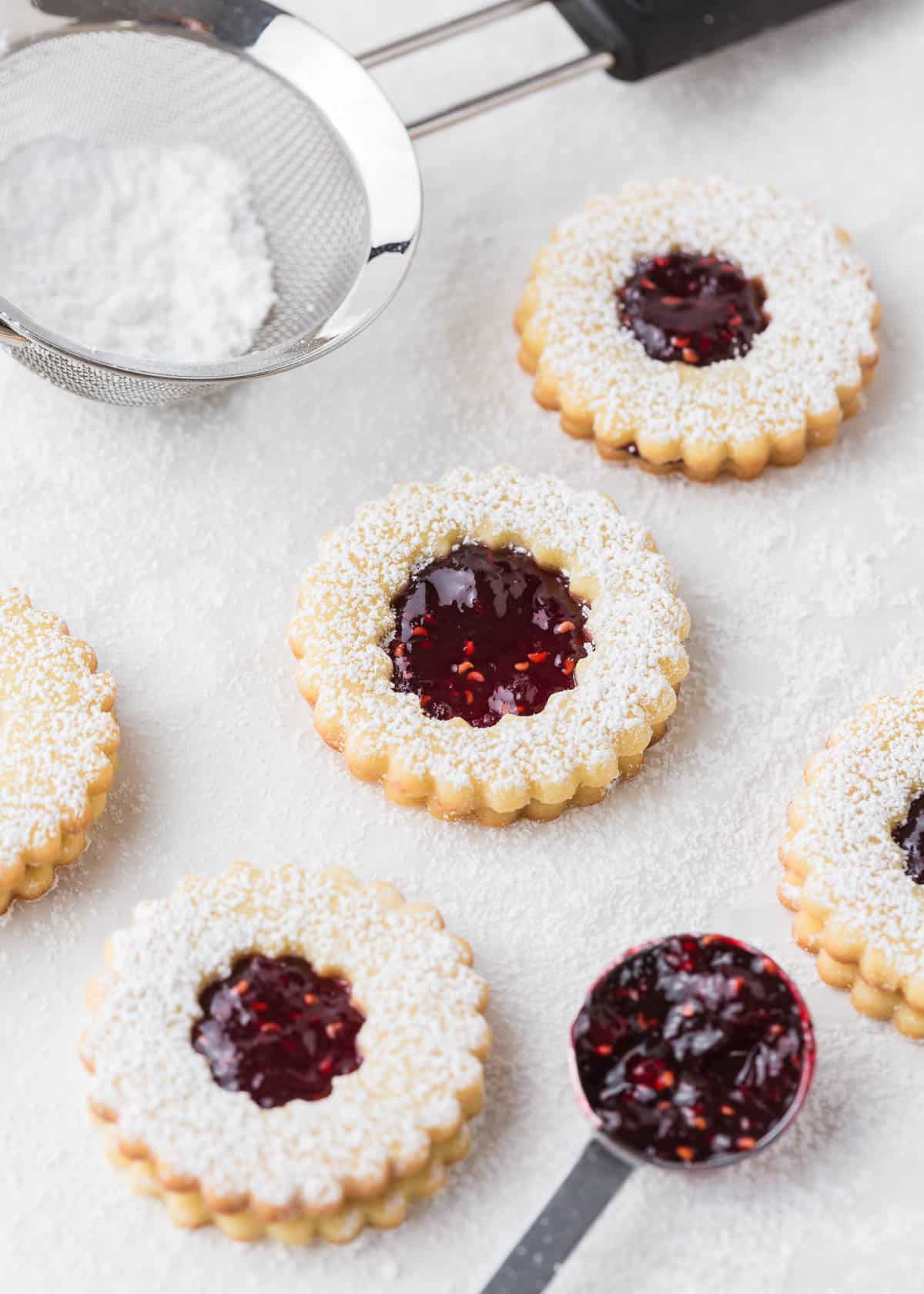 Linzer raspberry cookies on the counter with jam in a spoon and powdered sugar in a sifter.