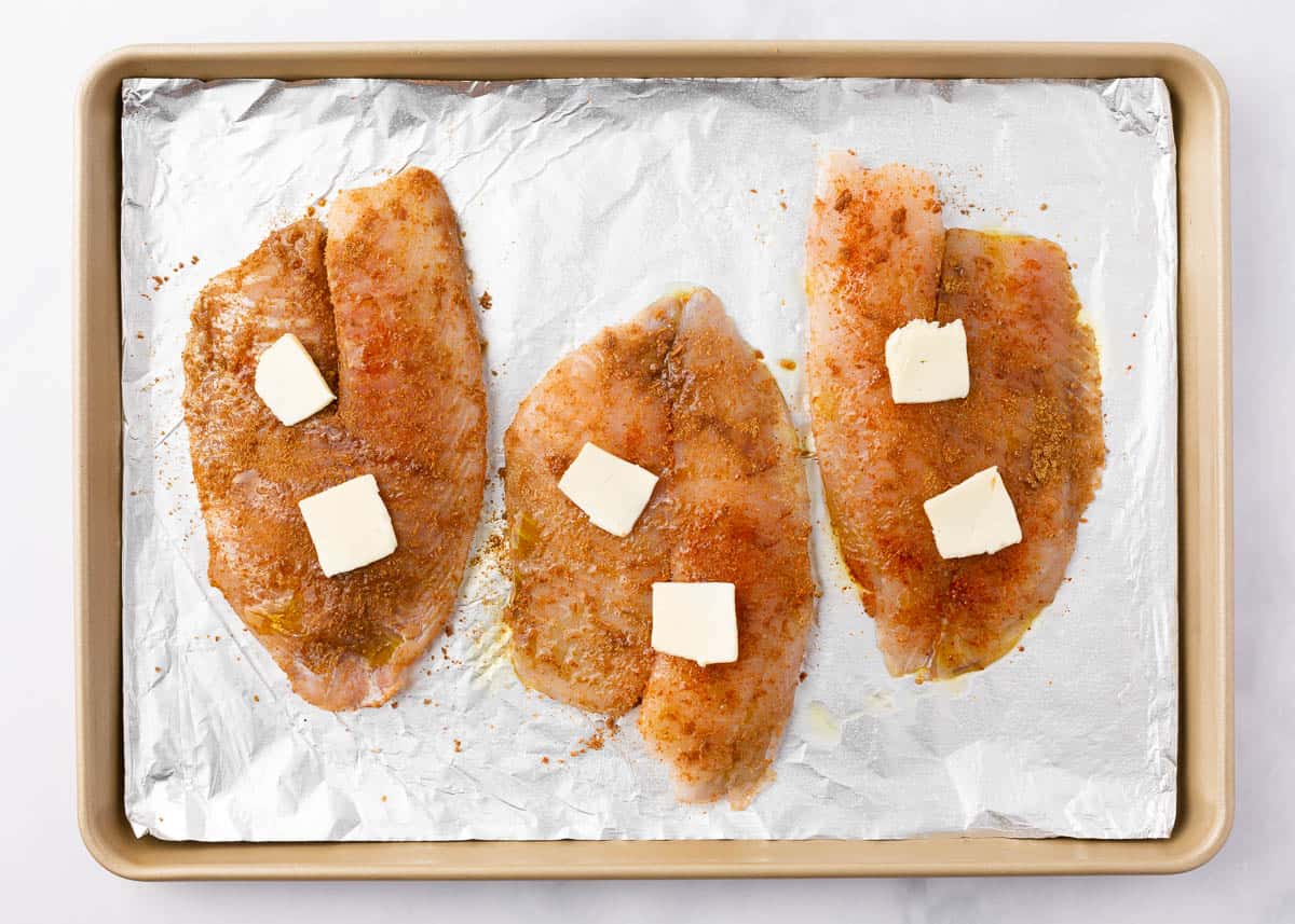 Tilapia on baking sheet with seasoning and pats of butter on each piece of fish.