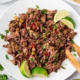 Barbacoa beef shredded on a plate with lime wedges.