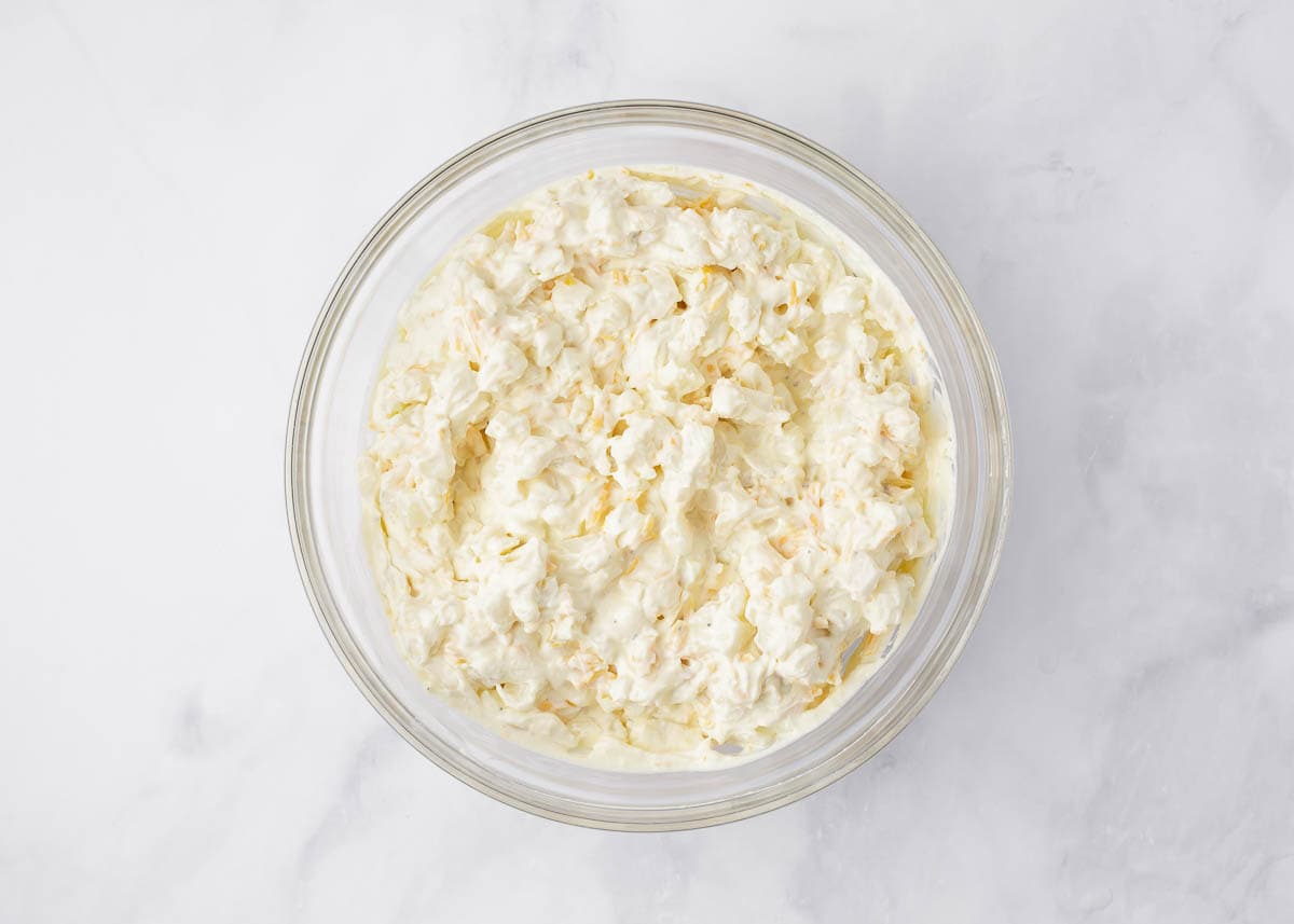 Potatoes and shredded cheese with creamy cheesy sauce mixture in a glass bowl.