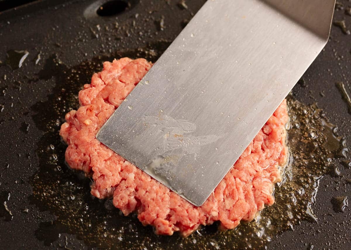 A burger being smashed with a metal spatula in a pan.