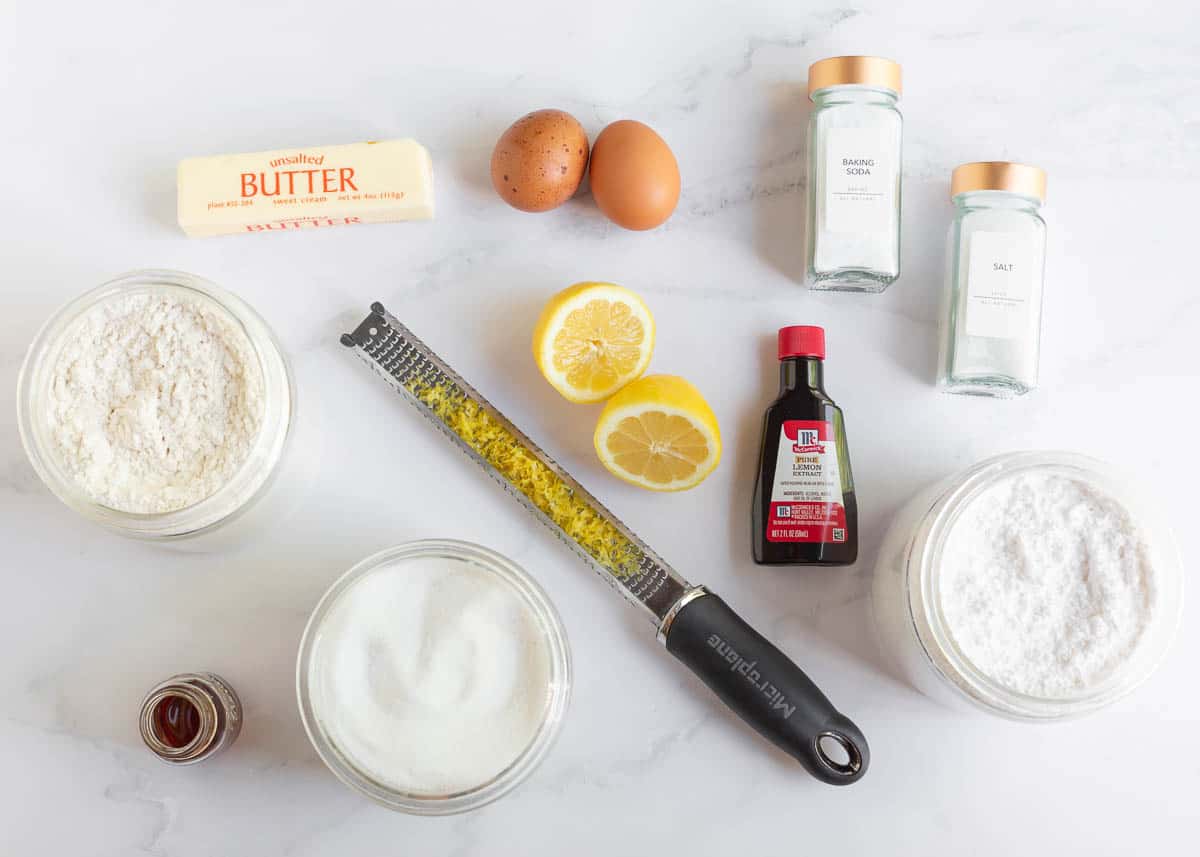 Lemon, lemon zest, powdered sugar, butter, eggs, flour, lemon extract, granulated sugar and baking staples on counter. 