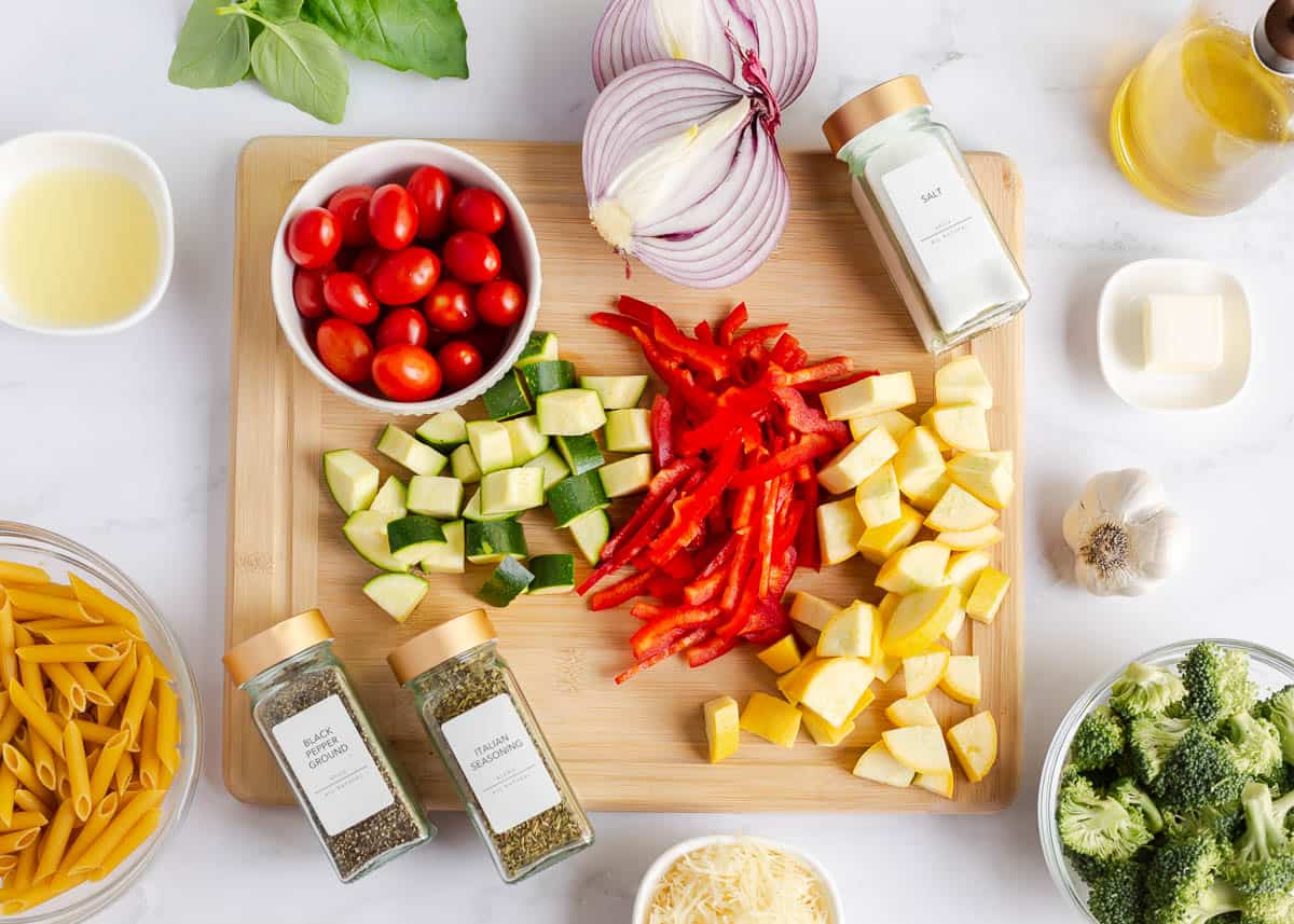 Penne pasta, tomatoes, summer squash, zucchini, red bell pepper, red onion, broccoli and seasonings on counter.