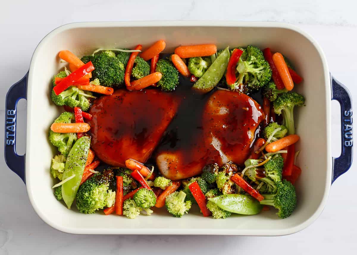 Vegetables with whole chicken pieces in a white baking dish with teriyaki sauce.