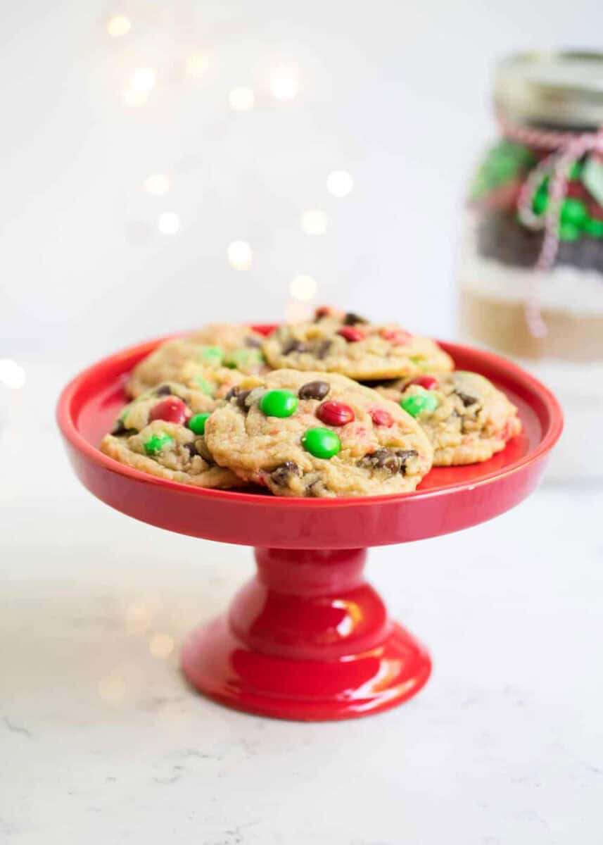 Homemade Christmas food gift idea, cookie mix in a jar with baked cookies on a red cookie stand.