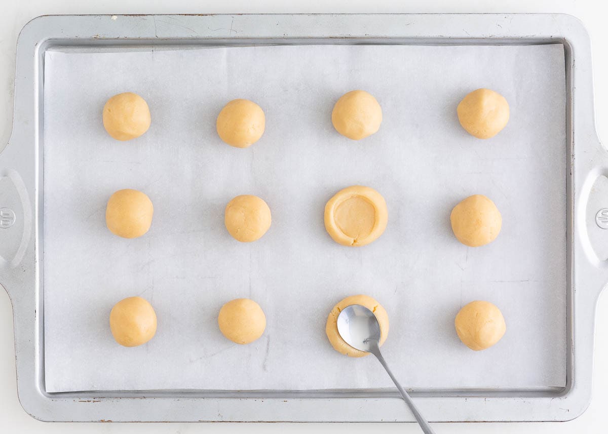 Thumbprint cookie dough on a baking sheet with a spoon making an indent in top of cookie.