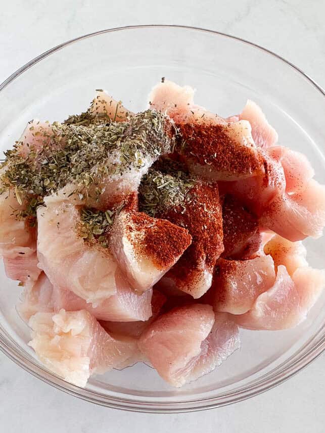 Raw chicken in bite sized pieces in a bowl with seasonings.